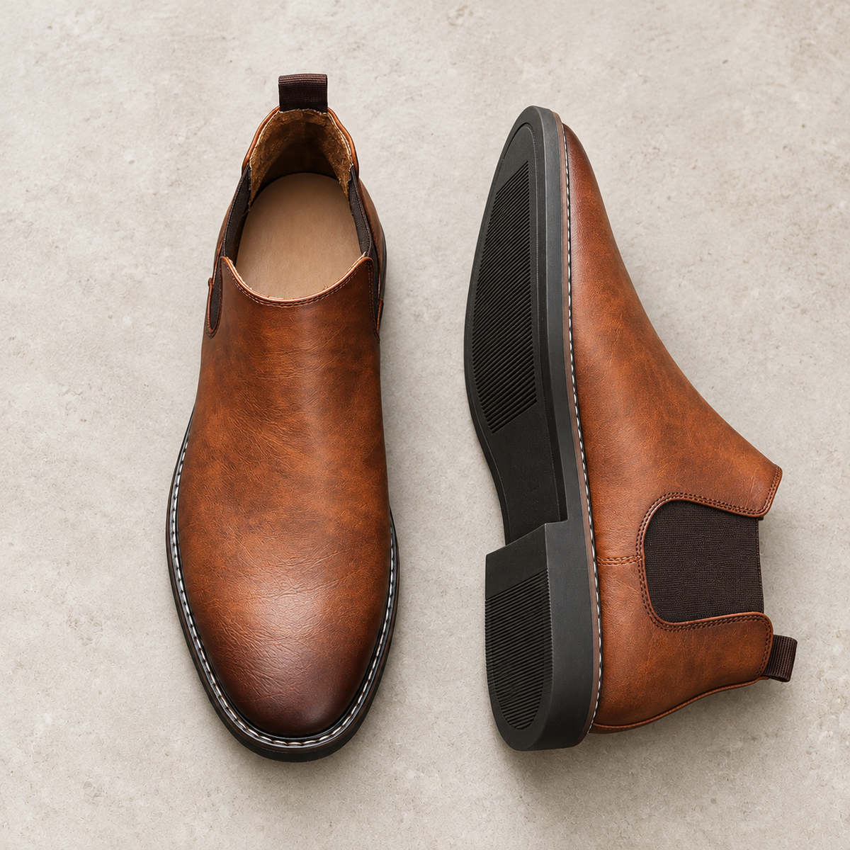 A pair of cognac brown Chelsea boots on pale limestone, showing the upper and black sole.